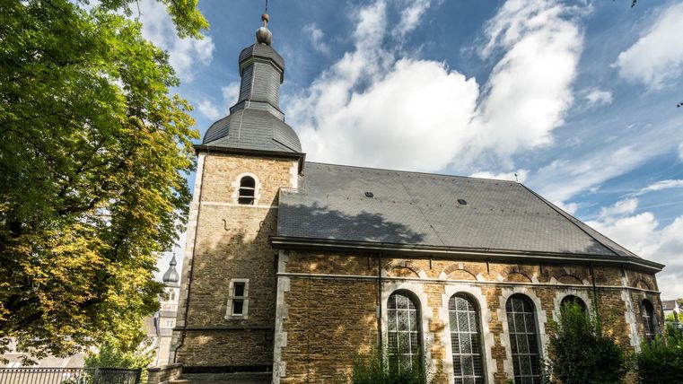The image shows a small church with a distinctive tower and large windows. Surrounded by green spaces and trees, it radiates a calm atmosphere.