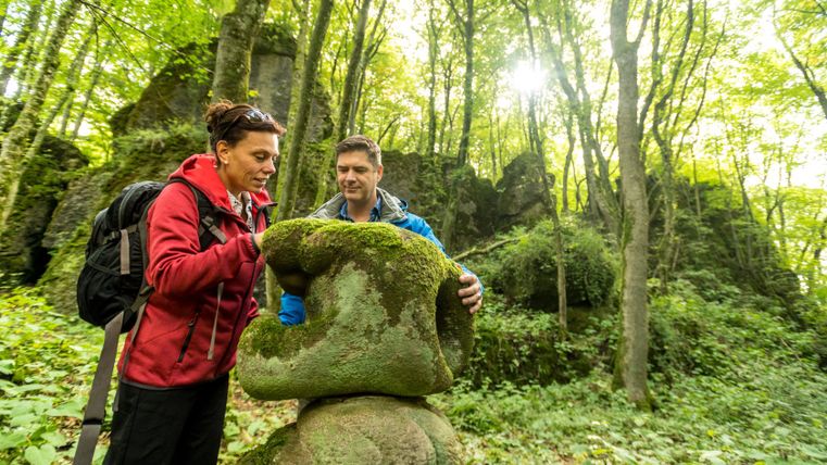 Zwei Personen betrachten einen moosbedeckten Stein im Wald.