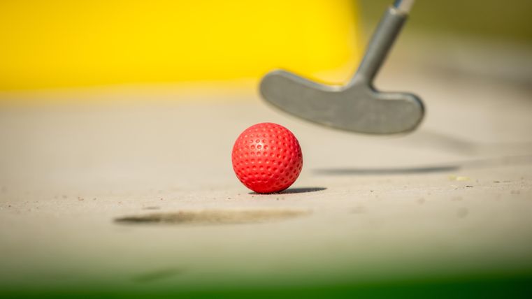 Ein roter Golfball liegt auf dem Boden. Ein Putter ist im Vordergrund sichtbar.