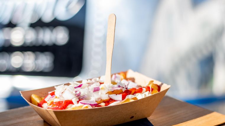 Een bord met friet, gegarneerd met verse ingrediënten zoals uien en tomaten. De eetkraam op de achtergrond is vaag te zien.