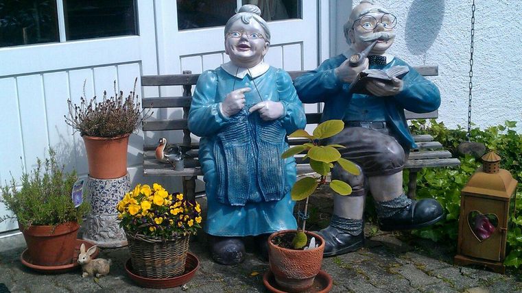 Zwei bunte Gartenfiguren sitzen auf einer Bank vor einem Haus. Sie sind mit Pflanzen und Töpfen umgeben, die eine freundliche Atmosphäre schaffen.