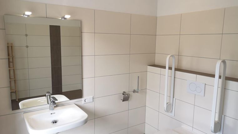 A modern bathroom with a bright tiled floor, a sink, and a mirror. Grab bars are installed on the wall for safety.