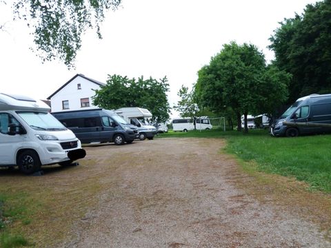 Wohnmobilstellplatz mit mehreren geparkten Wohnmobilen, umgeben von Bäumen und einem weißen Gebäude im Hintergrund.