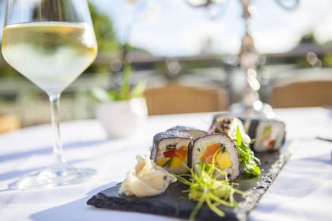 Een elegante tafel met sushirollen op een zwarte steen. Op de achtergrond staat een glas witte wijn en een klein bloempje.