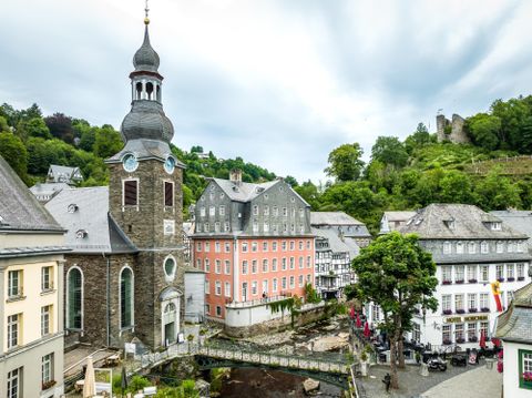 Ein malerischer Stadtplatz mit einer Kirche und historischen Gebäuden. Im Hintergrund ist ein Hügel mit Ruinen zu sehen.
