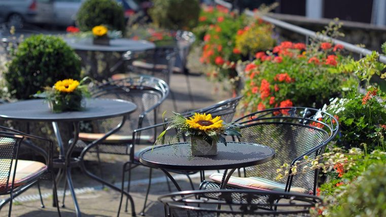Ein gemütlicher Außenbereich mit schwarzen Tischen und Stühlen. Auf den Tischen stehen kleine Pflanzen mit Sonnenblumen und umgeben von bunten Blumen.