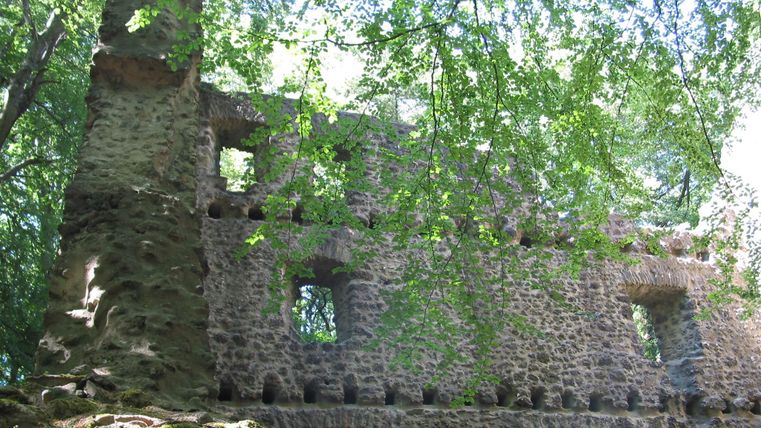 An old ruin, surrounded by trees and green foliage. The walls are partially at risk of collapse and show the marks of time.