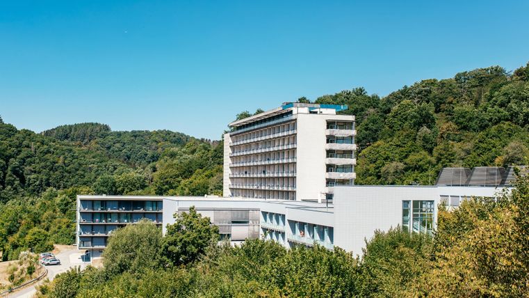 A modern hotel surrounded by green trees and hills. The clear sky offers a pleasant atmosphere.
