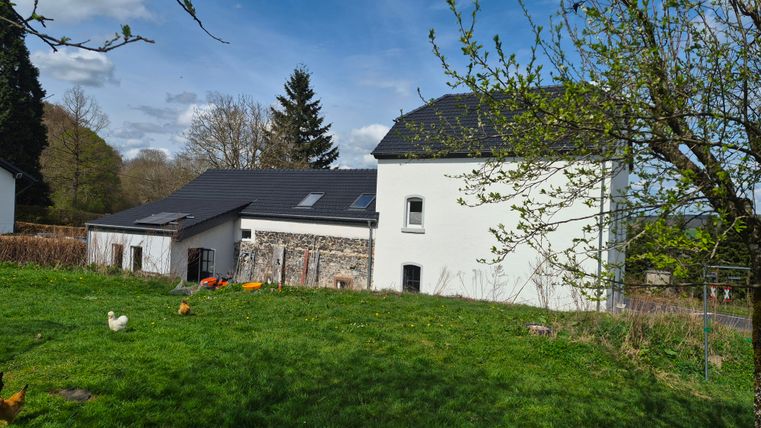 Ein großes, modernes Haus mit einem Walmdach und einer angelegten Wiese im Vordergrund. Im Hintergrund sind Bäume und ein klarer Himmel sichtbar.