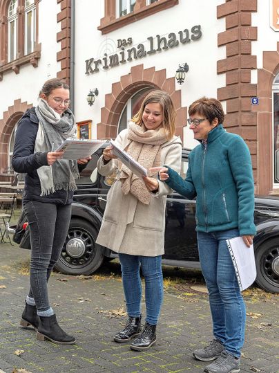 Three women are laughing in front of the crime house and looking at documents. In the background, an old car can be seen.