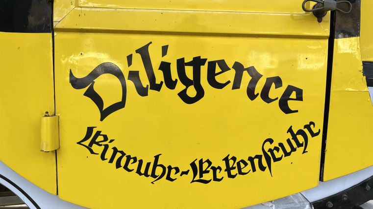 Een geel voertuig met het opschrift "Diligence". De tekst is in zwart, sierlijk lettertype geschreven.