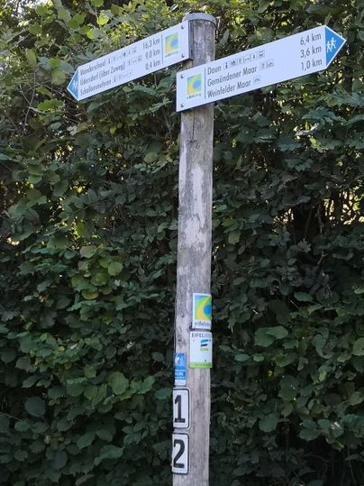 Ein Wegweiser mit mehreren Richtungspfeilen steht in einer grünen Umgebung. Die Schilder zeigen Entfernungen zu verschiedenen Zielen an.