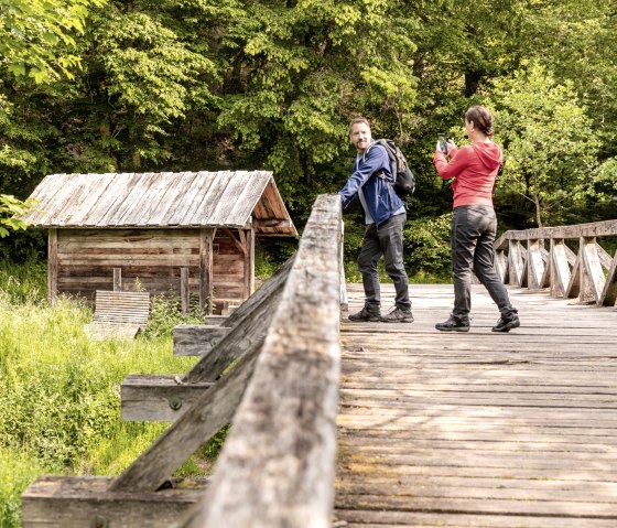 Zwei Personen stehen auf einer Holzbrücke vor einer kleinen Holzhütte, umgeben von üppigem Grün. Eine Person macht ein Foto., © Eifel Tourismus/AR-shapefruit AG