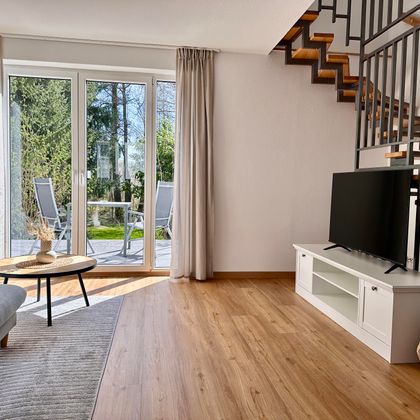 Ein helles Wohnzimmer mit einer gemütlichen Couch und einer modernen Treppe. Große Fenster lassen viel Licht herein und bieten einen Blick auf den Außenbereich.