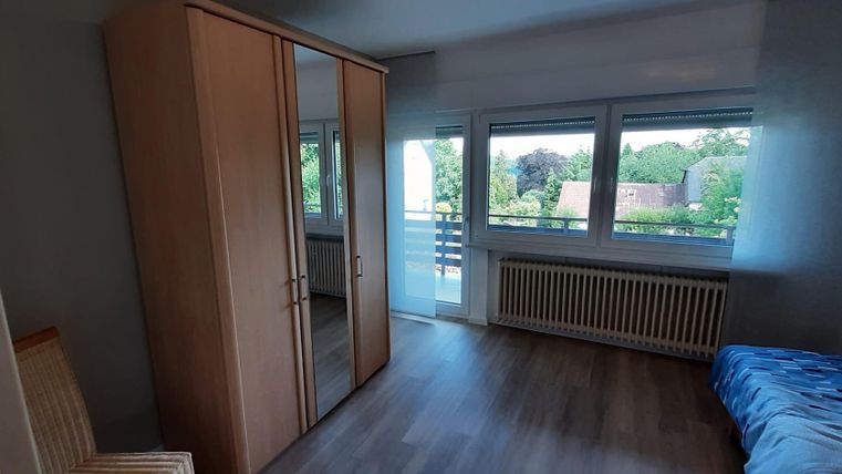 Ein helles Zimmer mit großen Fenstern und Blick ins Freie. Die Einrichtung umfasst einen Kleiderschrank und ein Bett mit einer blauen Bettdecke.