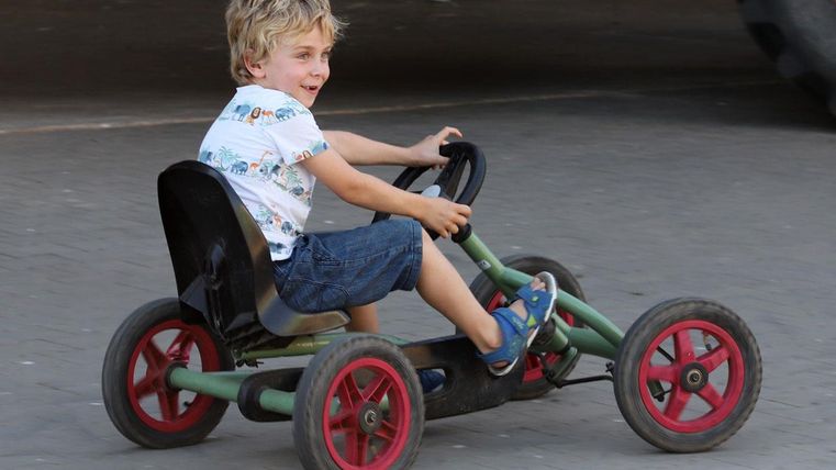 A boy is happily riding on a go-kart. The surroundings appear to be a playground or recreational area.