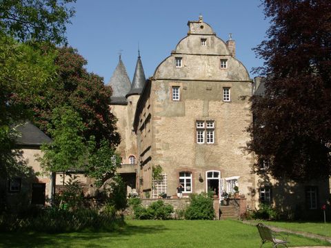 Historisches Gebäude mit Türmen, umgeben von Bäumen und einem gepflegten Garten. Eine Terrasse ist sichtbar, das Wetter ist sonnig.