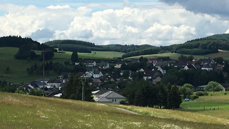 Eine malerische Landschaft mit sanften Hügeln und einem kleinen Dorf im Hintergrund. Der Himmel ist teils bewölkt mit einigen Sonnenstrahlen.