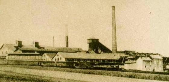 An old factory complex with tall chimneys and several buildings. The surroundings are rural and expansive.