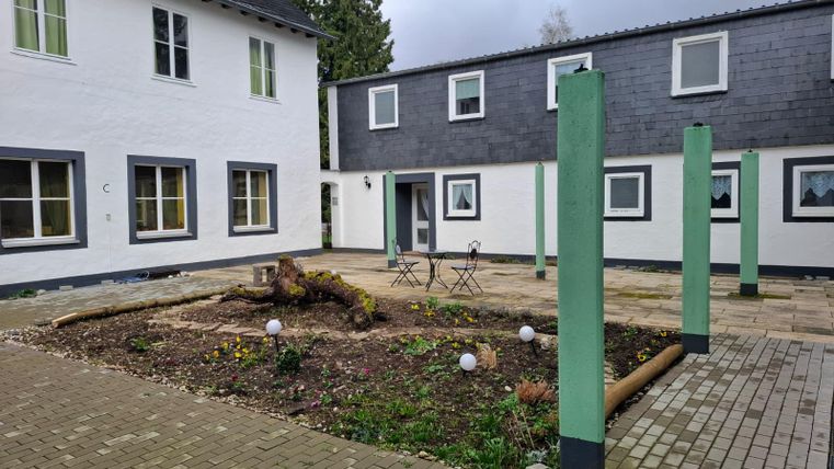 A small courtyard with two buildings and a green area in the middle. There are some chairs and columns as well as decorative spheres.