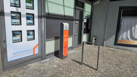 Ein moderner Parkplatz mit einem Fahrradständer und einem Informationsstand. Die Fassade des Gebäudes zeigt Werbefenster mit Bildern.