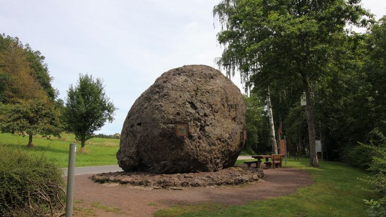 Een grote, ronde rots staat in een groene omgeving. Ernaast staat een bank en bomen op de achtergrond.