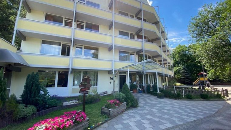 A modern building with several floors surrounded by green trees and colorful flower beds. The entrance area is welcoming and appears friendly.