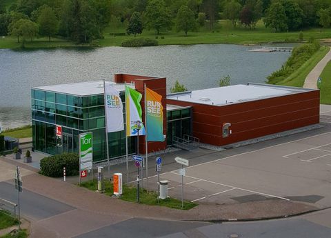 Ein modernes Gebäude am Wasser mit großen Fenstern. Im Vordergrund sind Flaggen und eine gepflasterte Fläche zu sehen.