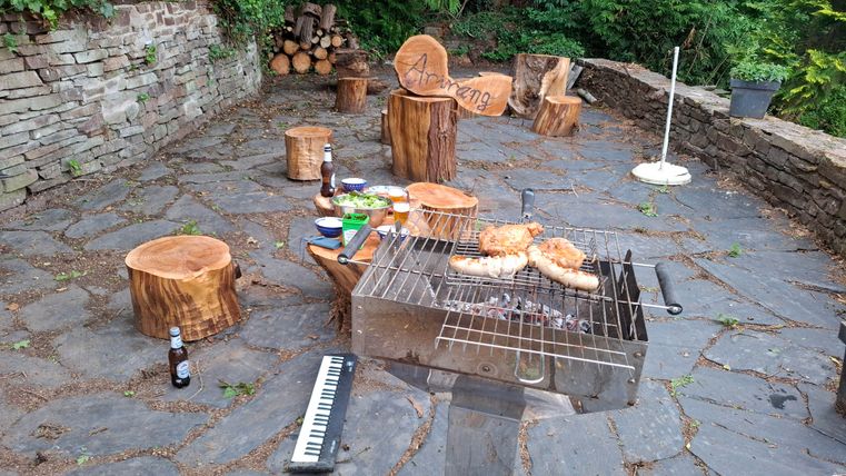 A rustic outdoor grill area with log seats. Grilled sausages are on the grill and some drinks are scattered on the ground.