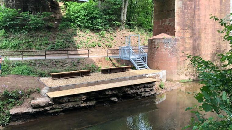 Ein ruhiger Wasserbereich unter einer Brücke, umgeben von grünem Bewuchs. Eine kleine Holzbank lädt zum Verweilen ein.