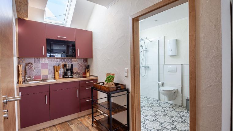 Eine moderne Küche mit dunklen Schränken und einem offenen Badezimmer. Helle Fliesen und ausreichend Tageslicht schaffen eine einladende Atmosphäre.