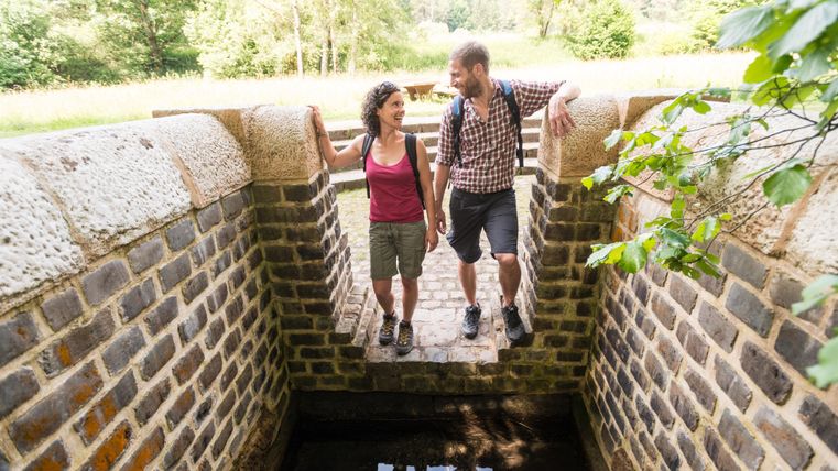 A couple stands on a small stone step above a waterway in a green setting. In the background, trees and grass can be seen.