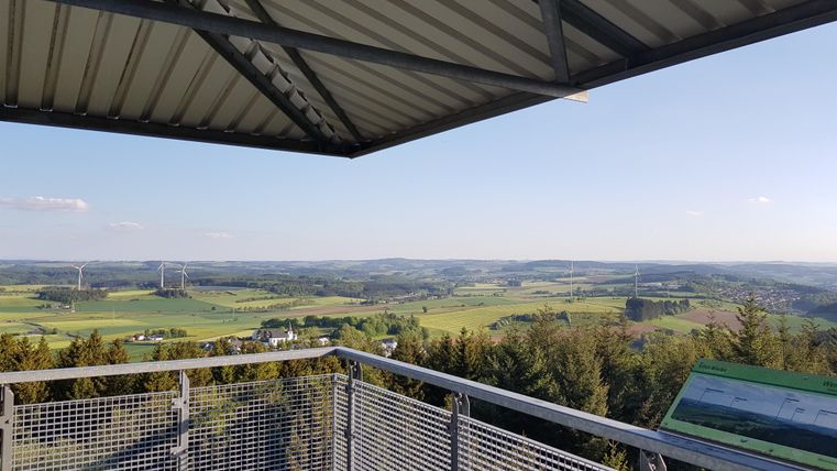 A view from an observation platform shows a green landscape with gentle hills and wind turbines in the background. The sky is clear and blue, providing a beautiful view.
