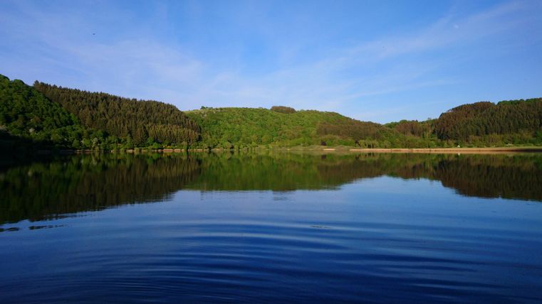 Een rustige meer, omgeven door groene heuvels en bossen. Het water weerspiegelt de heldere blauwe lucht.