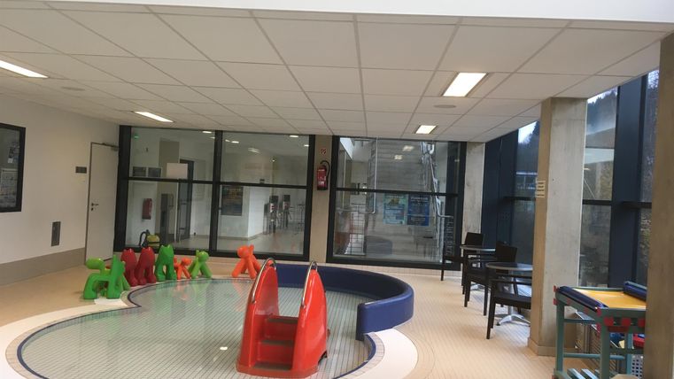 Een lichte ruimte met een klein kindergebied dat een ondiep waterbasin en gekleurde speeltoestellen bevat. Er zijn grote ramen die veel daglicht binnenlaten.