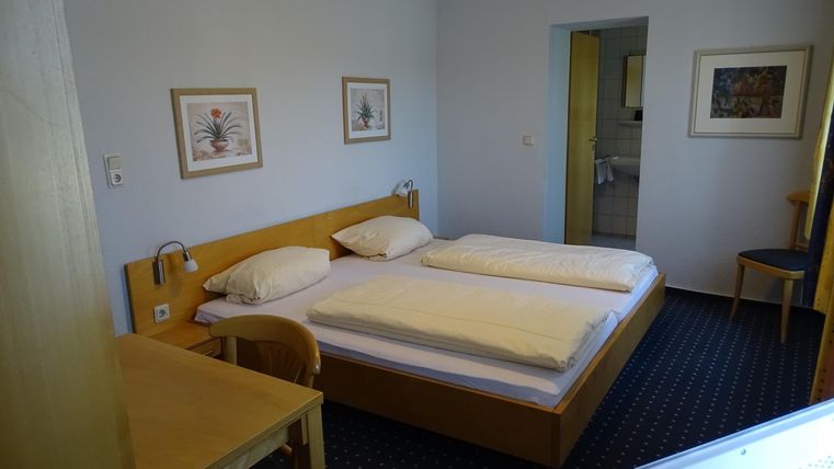 Ein einfaches Gästezimmer mit zwei Einzelbetten und einem Tisch. Die Wände sind mit Bildern dekoriert, und es gibt einen Blick auf ein Badezimmer.