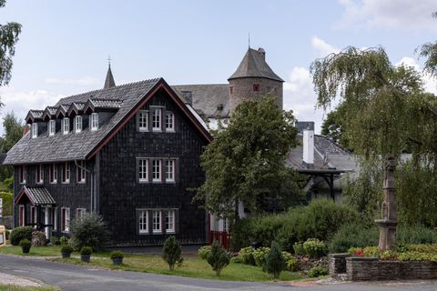 Ein charmantes Gebäude mit dunklen Schindeln und einem Turm. Es ist von üppigem Grün umgeben und hat eine einladende Atmosphäre.
