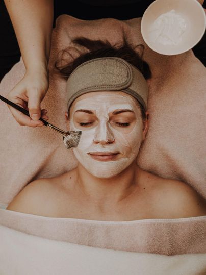 Eine Frau erhält eine Gesichtsbehandlung mit einer Maske. Sie liegt entspannt auf einem Handtuch, während ein Behandler die Maske aufträgt.
