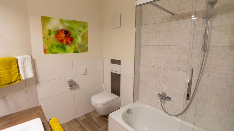 Ein modernes Badezimmer mit einer Badewanne und einer Dusche. An der Wand hängt ein buntes Bild einer Blume und es gibt gelbe Handtücher.