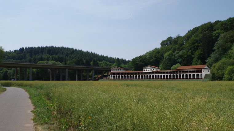 Ein ruhiges Feld mit sanften Hügeln im Hintergrund. Im Vordergrund steht ein Weg und im Hintergrund ist ein historisches Gebäude zu sehen.