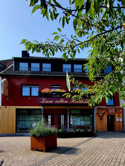 Ein gemütliches Gasthaus mit roter Fassade und einer einladenden Terrasse. Umgeben von Bäumen und einem gepflasterten Platz.