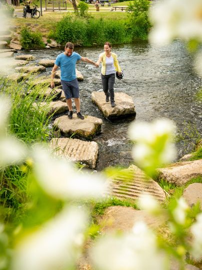 Een paar steekt op stenen een rivier over in het Kurpark Gerolstein. Op de voorgrond zijn onscherpe bloemen, op de achtergrond groene weilanden en bomen.