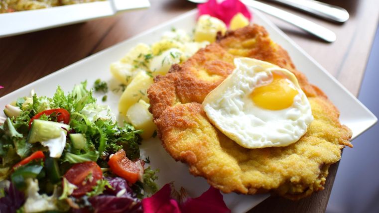 Een bord met een schnitzel, daarop een spiegelei, daarnaast aardappelen en een gemengde salade. Op de achtergrond bestek.