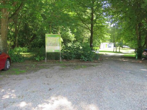Een smal grindpad tussen bomen leidt naar een parkeerplaats. Op de achtergrond zijn nog meer bomen en een bord zichtbaar.