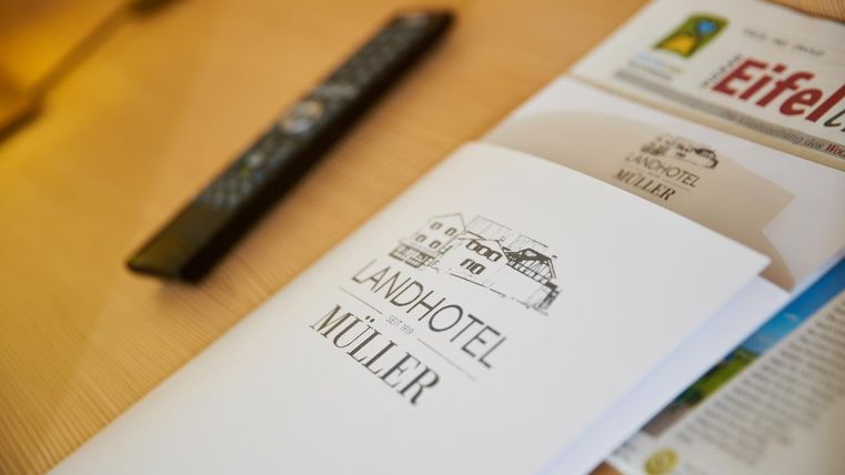 Ein offenes Faltblatt des Landhotels Müller liegt auf einem Tisch. Daneben befindet sich eine Fernbedienung und eine Zeitschrift über die Eifel.