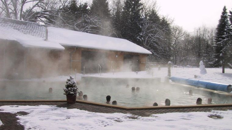 A heated outdoor swimming pool, surrounded by snow and steam. In the background, there is a wooden building and a few trees are visible.