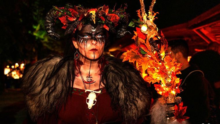 Een vrouw in een opvallende kostuum met een hoofddeksel van hoorns en kleurrijke bladeren. Ze houdt een stralend accessoire vast en heeft een dramatische make-up.