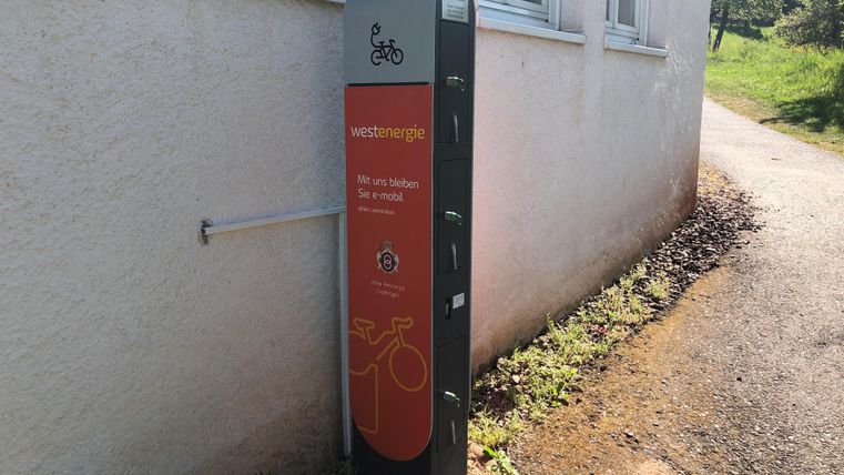 Eine Fahrradstation mit einem orangefarbenen Schild und verschiedenen Fächern. Sie befindet sich an einer Wand, umgeben von Gras und einem Schotterweg.