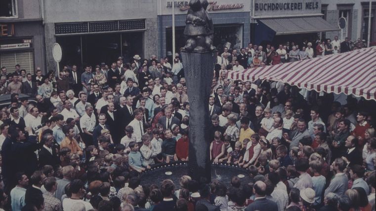 Een grote menigte verzamelt zich rond een fontein met een standbeeld in het midden. Op de achtergrond zijn luifels en winkels te zien.