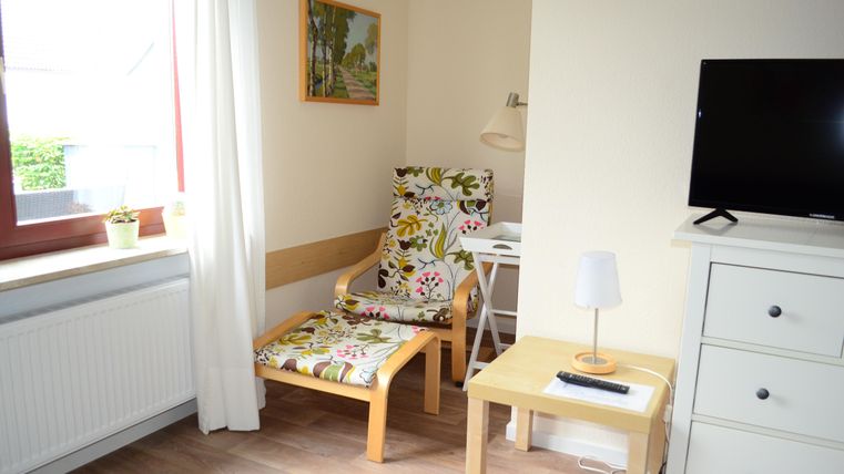 A cozy living area with a comfortable chair and a small table. There is a painting on the wall, and a TV is placed on a dresser.